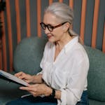 Elderly woman in glasses reviewing documents on clipboard, sitting indoors with a focused expression.