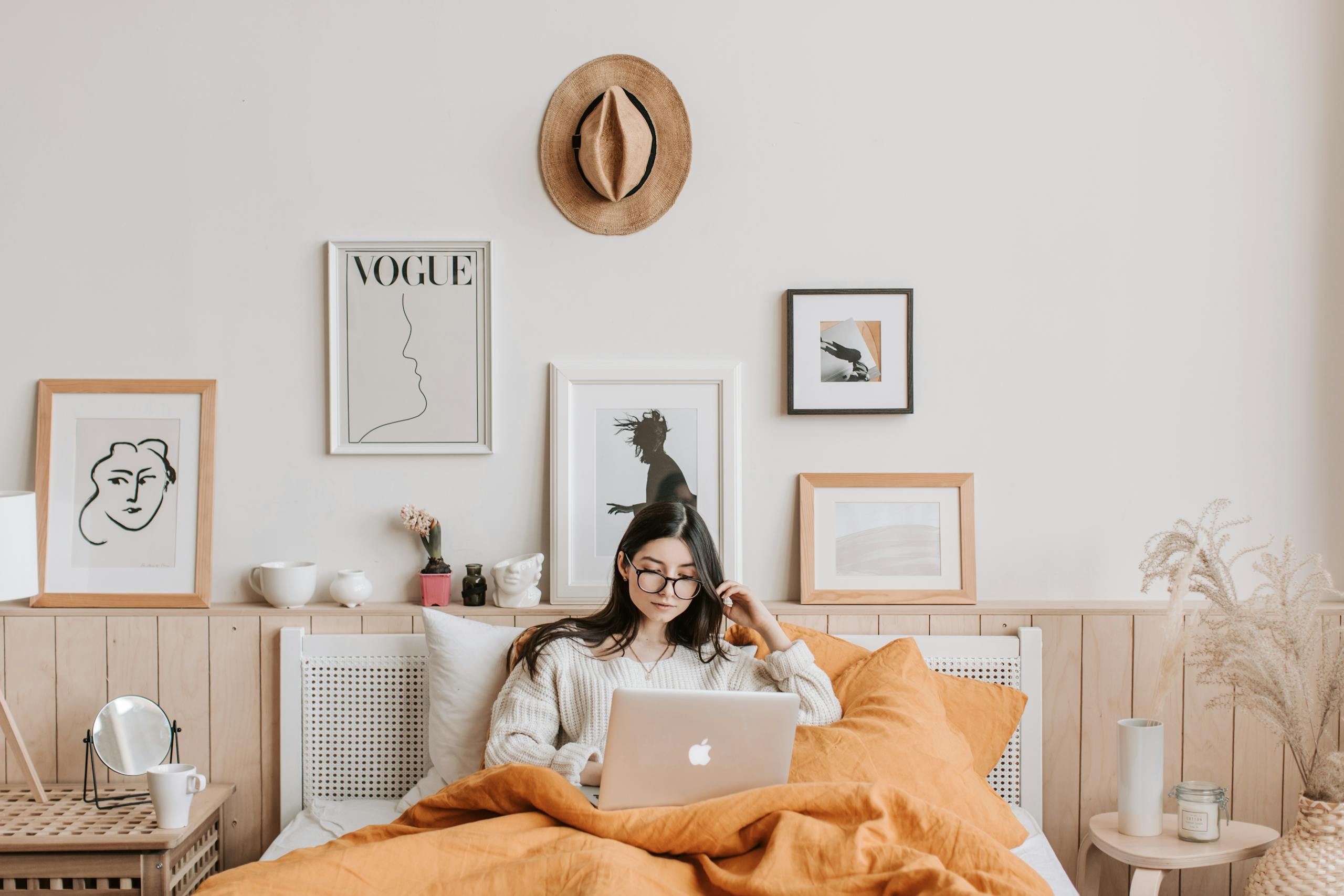 Young woman in cozy bedroom using a laptop, surrounded by stylish decor and art.